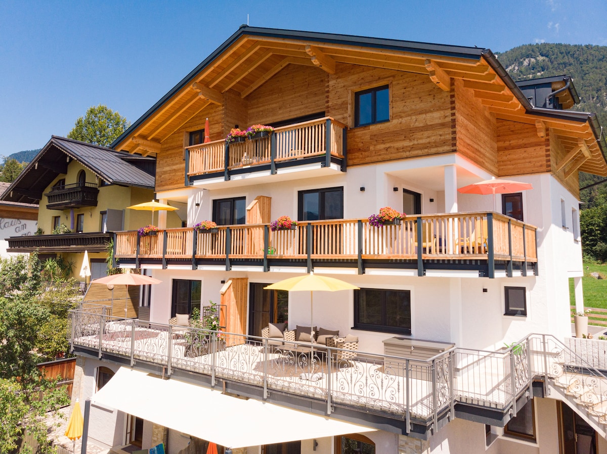 The exterior of a multi-level accommodation features wooden balconies adorned with colorful flowers. Sun umbrellas provide shade over patio seating on the ground floor, while large windows allow natural light into the interior. Surrounding mountains are visible in the background.