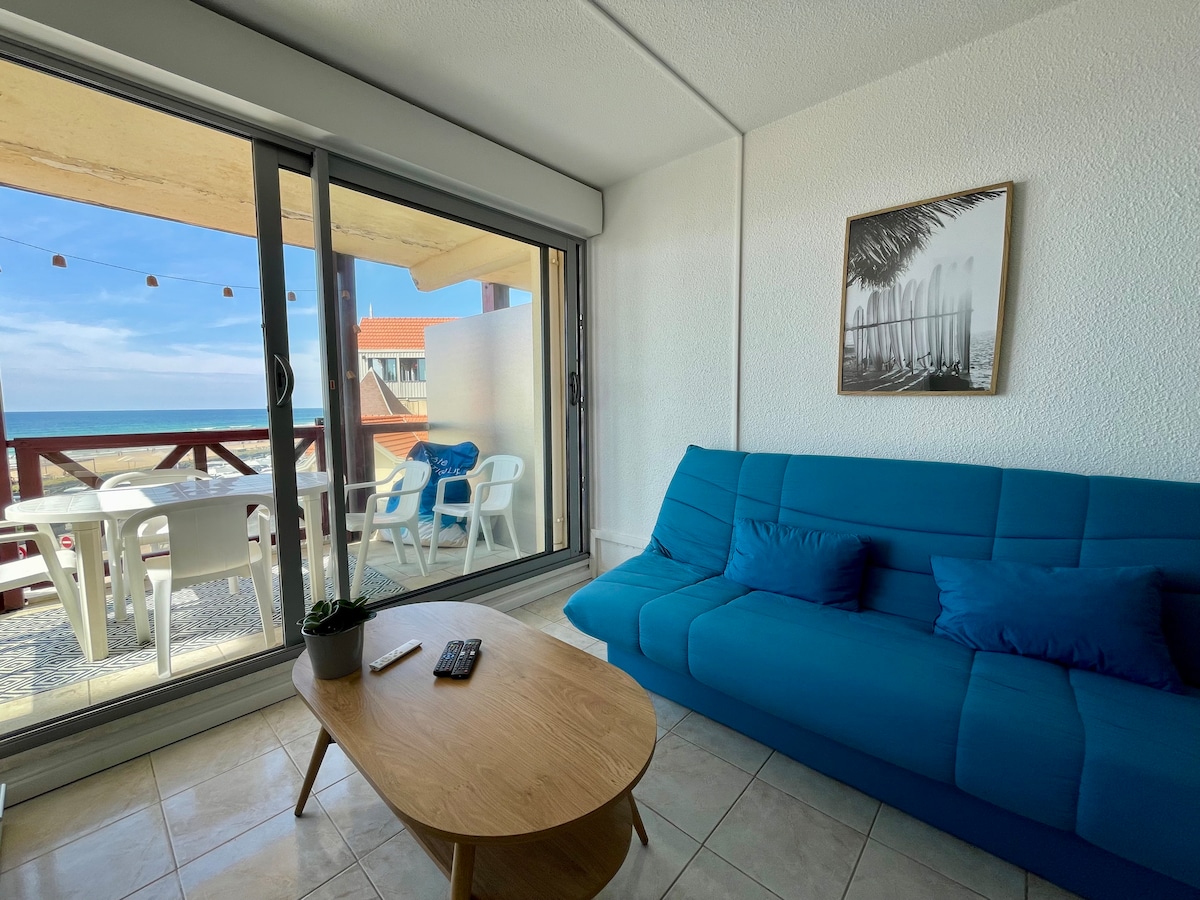 A light-filled living space features a blue sofa and a wooden coffee table. Large glass doors open to the terrace, showcasing a view of the ocean. A dining set with white chairs is visible on the terrace, enhancing indoor-outdoor connectivity.