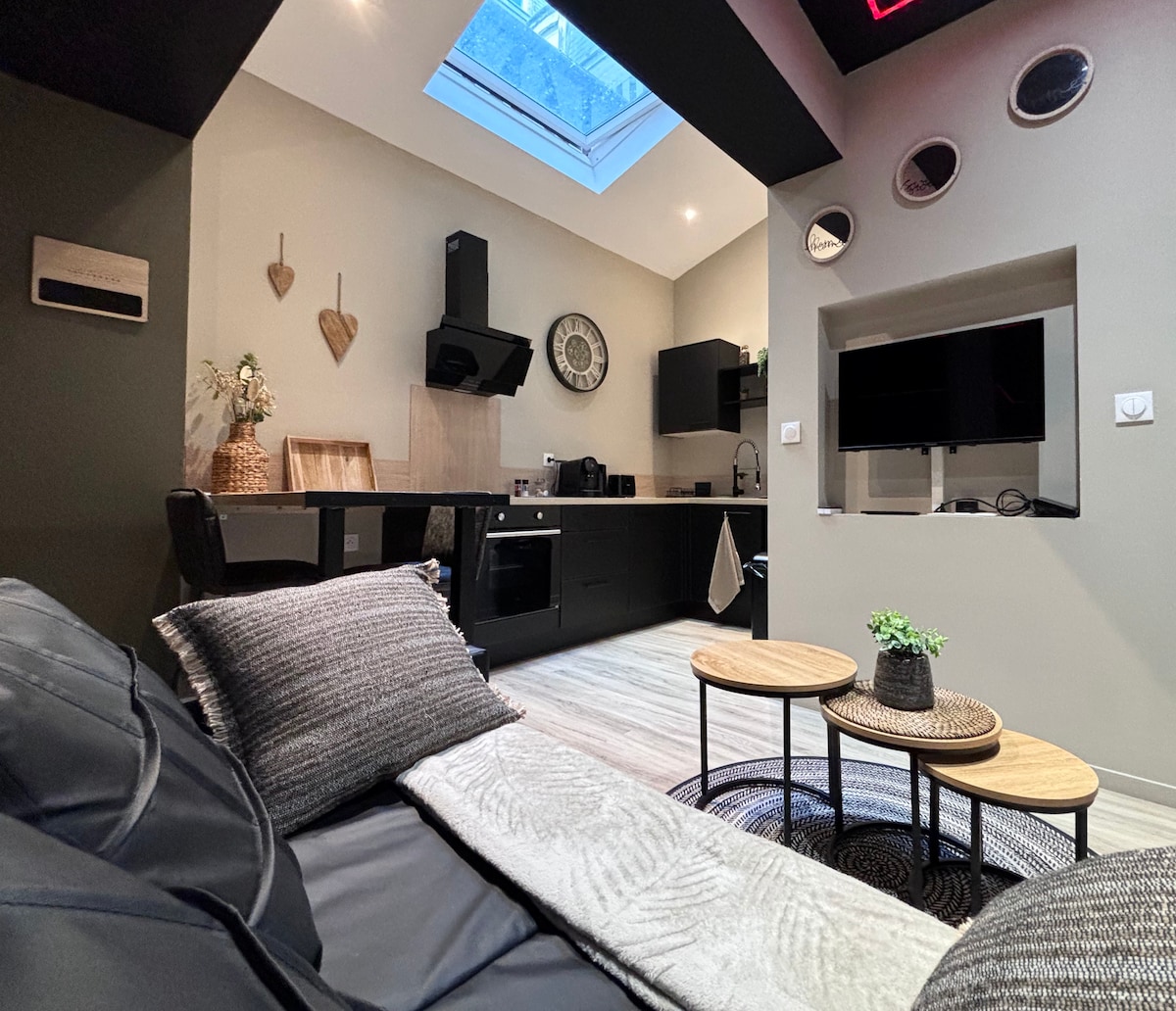 A modern kitchen area features sleek black cabinetry and countertop appliances, complemented by a decorative wall clock. Natural light pours in through a skylight, illuminating a cozy seating space with a textured couch and two round coffee tables adorned with a plant. Wall mirrors enhance the ambiance.