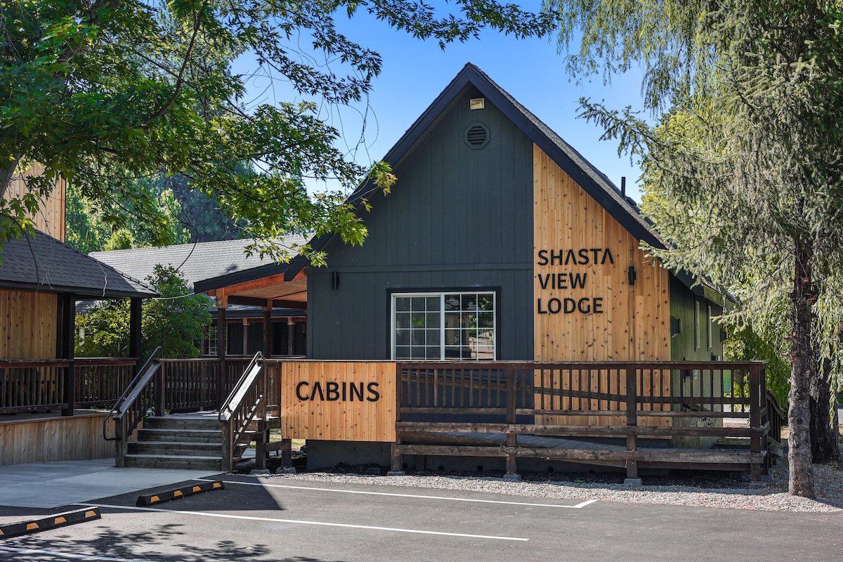 The exterior of Shasta View Lodge is showcased, featuring a combination of dark and light wood siding. A large front porch with a wooden railing extends across the entrance. Lush trees frame the building, enhancing the welcoming atmosphere.