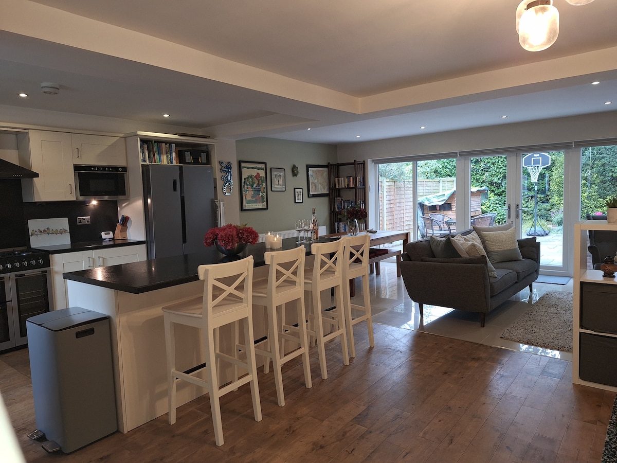 A modern kitchen and living area is featured, showcasing a sleek kitchen island with four high chairs. Light-colored cabinetry complements the stainless steel appliances. A cozy seating arrangement with a plush sofa is positioned near glass doors leading to an outdoor space, illuminated by natural light.