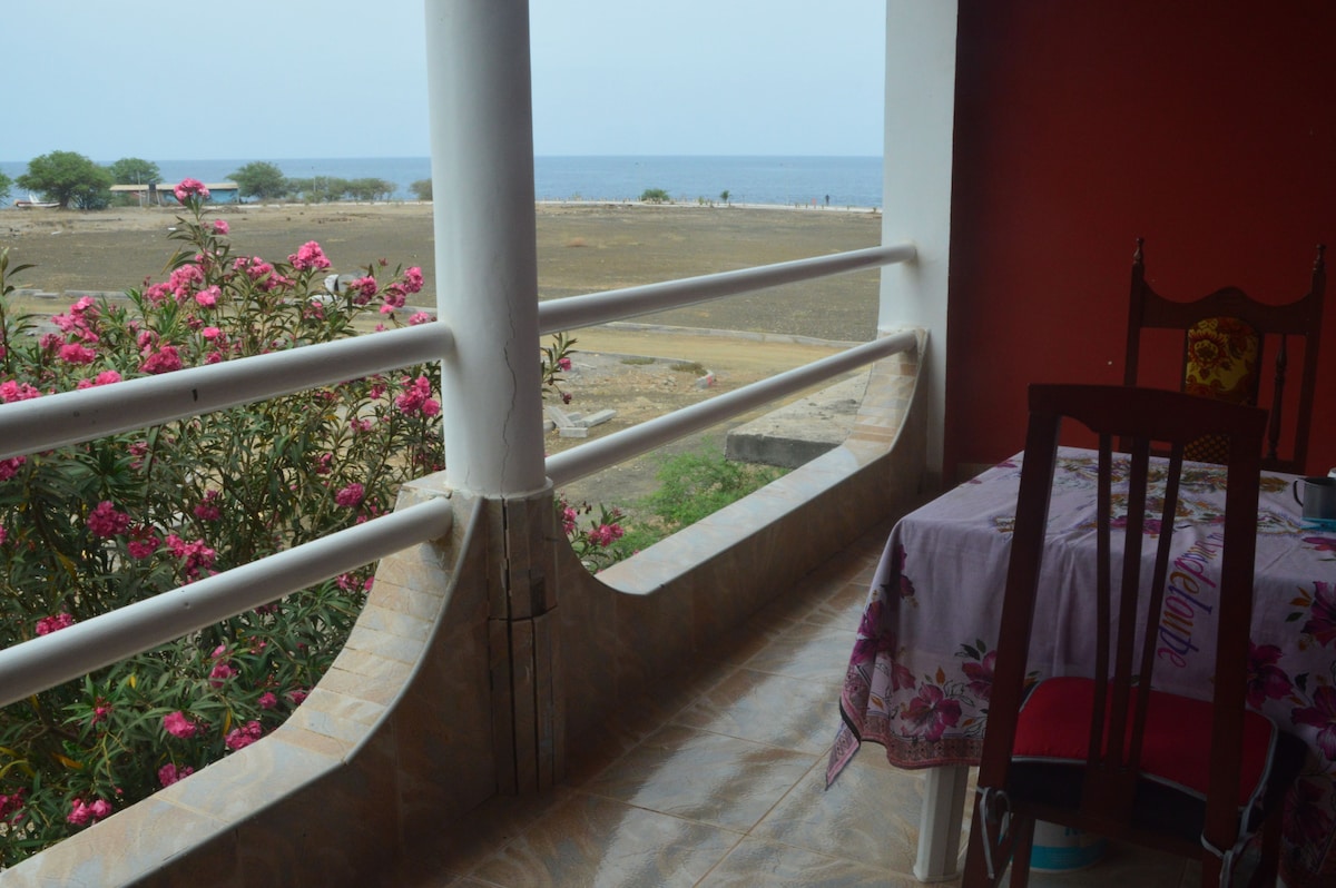 A covered patio offers a view of the sea, framed by flowering shrubs and trees. A dining table with four wooden chairs sits on the patio, with a colorful tablecloth draped over it. The peaceful setting promotes relaxation and enjoyment of the ocean breeze.
