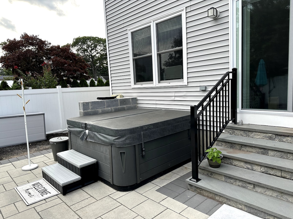 An outdoor hot tub, covered, is positioned beside a set of stairs leading to a spacious deck area. The area is paved with large tiles, and a small potted plant rests near the base of the stairs. A white fence and trees are visible in the background.