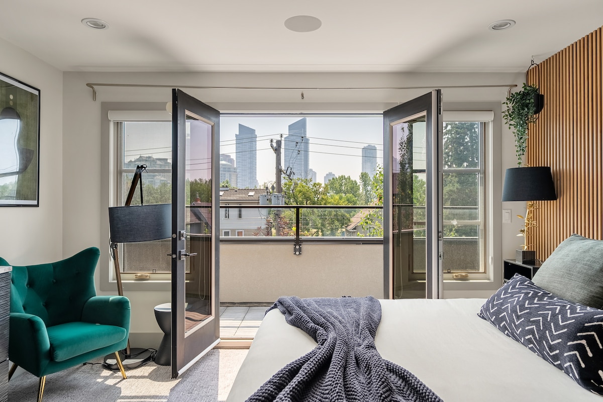 The master bedroom features a spacious layout with two sets of French doors that open onto a private balcony. A beautiful view of downtown Calgary's skyline is visible in the background. A comfortable chair and lamp are positioned beside the bed, adding functionality to the space.
