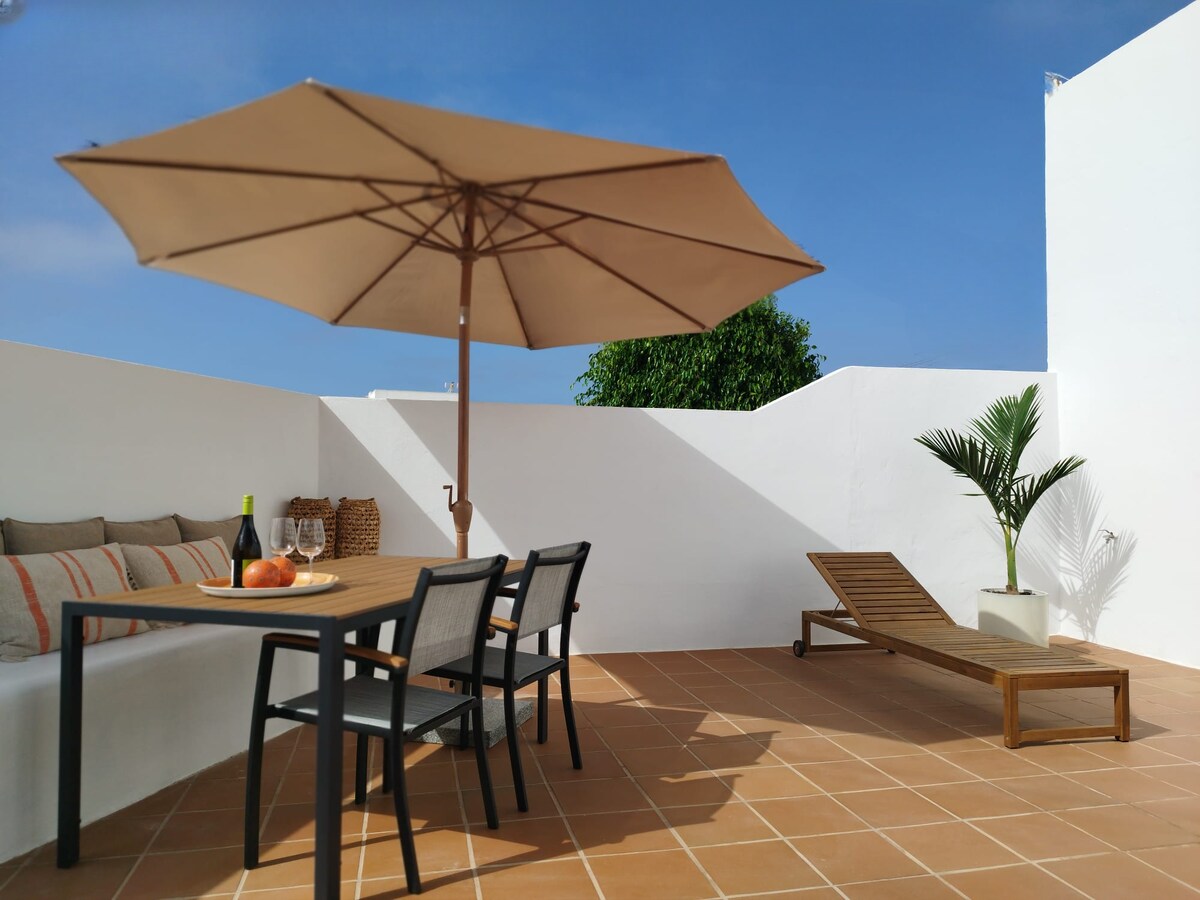 A spacious terrace features a wooden dining table set for two, accompanied by a large parasol providing shade. A comfortable lounge chair and a decorative plant are positioned nearby. Soft seating with cushions is also available against the wall, inviting relaxation.