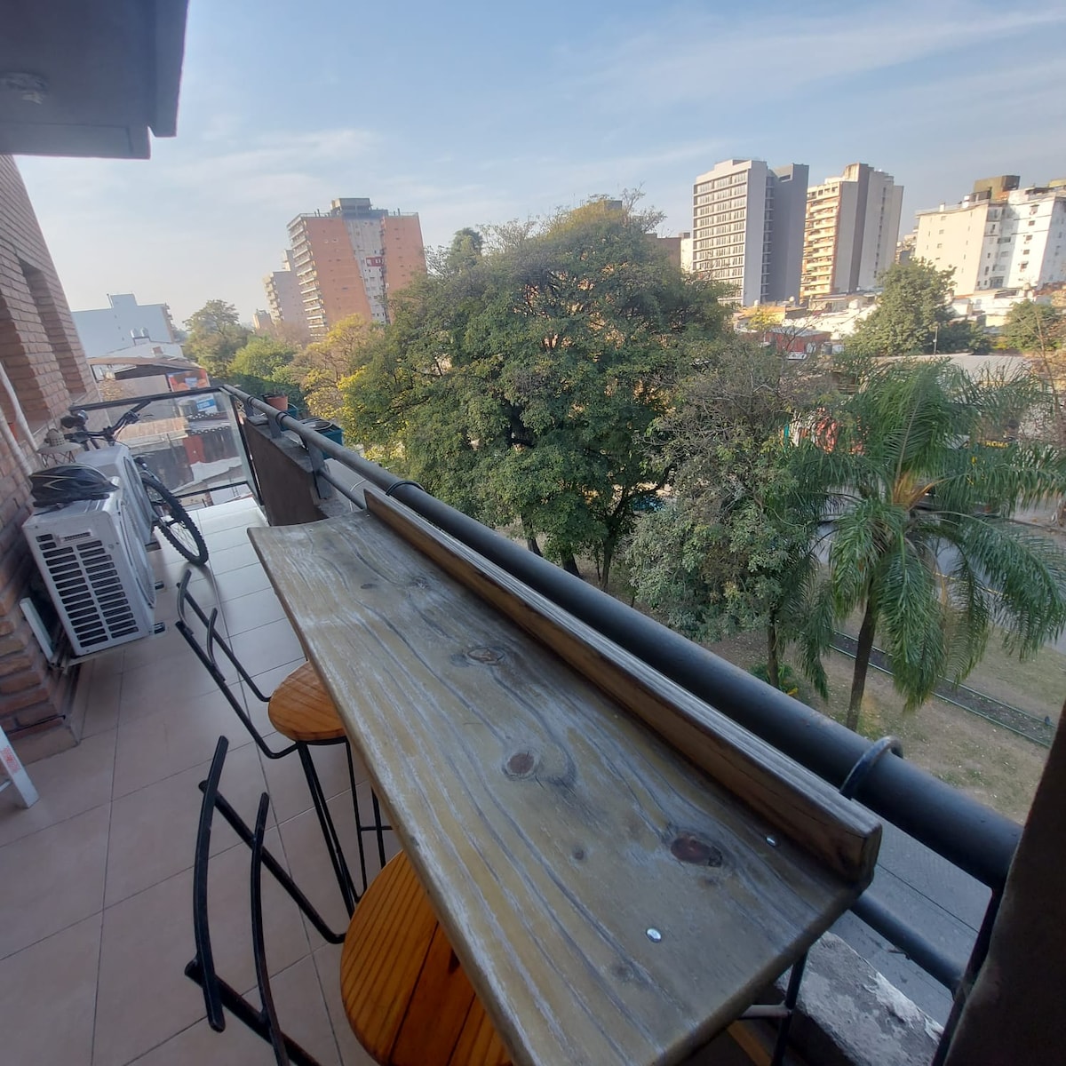A balcony area features a wooden bar table accompanied by several round stools, providing a seating space to enjoy the outdoors. Lush trees and buildings are visible in the distance, and an air conditioning unit can be seen mounted on the wall.
