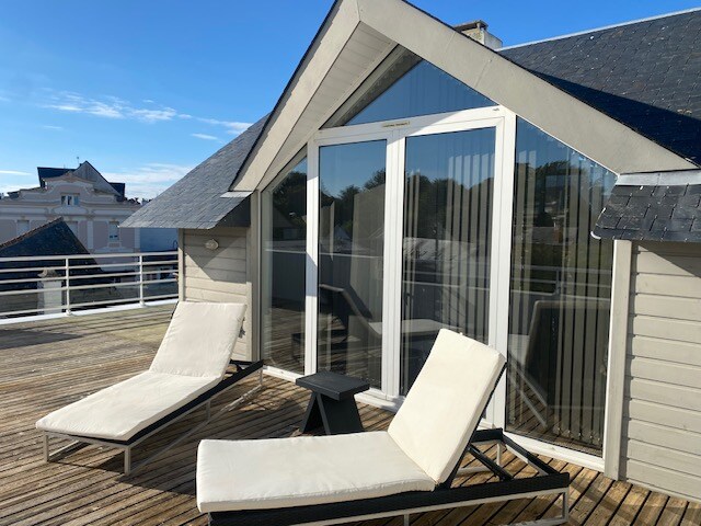 The rooftop terrace features two lounge chairs positioned on a wooden deck, overlooking the landscape. Large glass doors provide access to the interior, allowing natural light to illuminate the space. The sloped roof adds an architectural detail, enhancing the overall design.