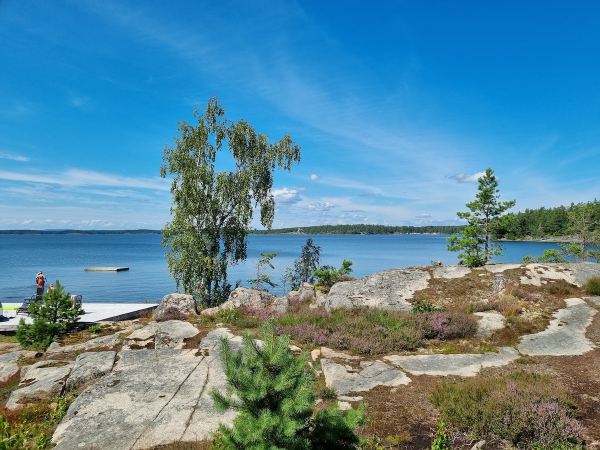 A serene lakeside view features calm, blue waters under a clear sky. Rocky terrain with patches of greenery and vibrant wildflowers is visible. In the distance, a wooden dock extends into the lake, surrounded by trees, creating a peaceful outdoor setting.