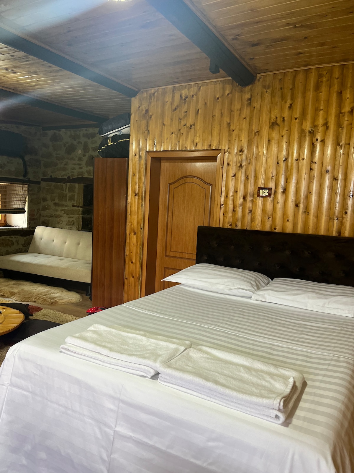 A sleeping area features a spacious bed with white bedding, accompanied by neatly folded towels. The wooden walls and ceiling add warmth to the space. A seating area with a light-colored couch can be seen in the background, creating a cozy atmosphere.