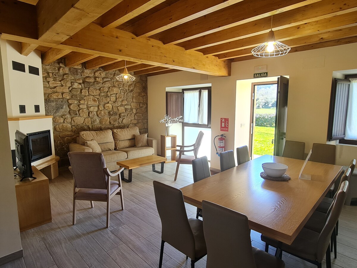 A spacious living area is presented with a wooden ceiling and light-colored walls. A comfortable sofa is positioned near a stone feature wall, while a large dining table is visible, surrounded by several chairs. Natural light enters through the windows, creating a bright atmosphere.