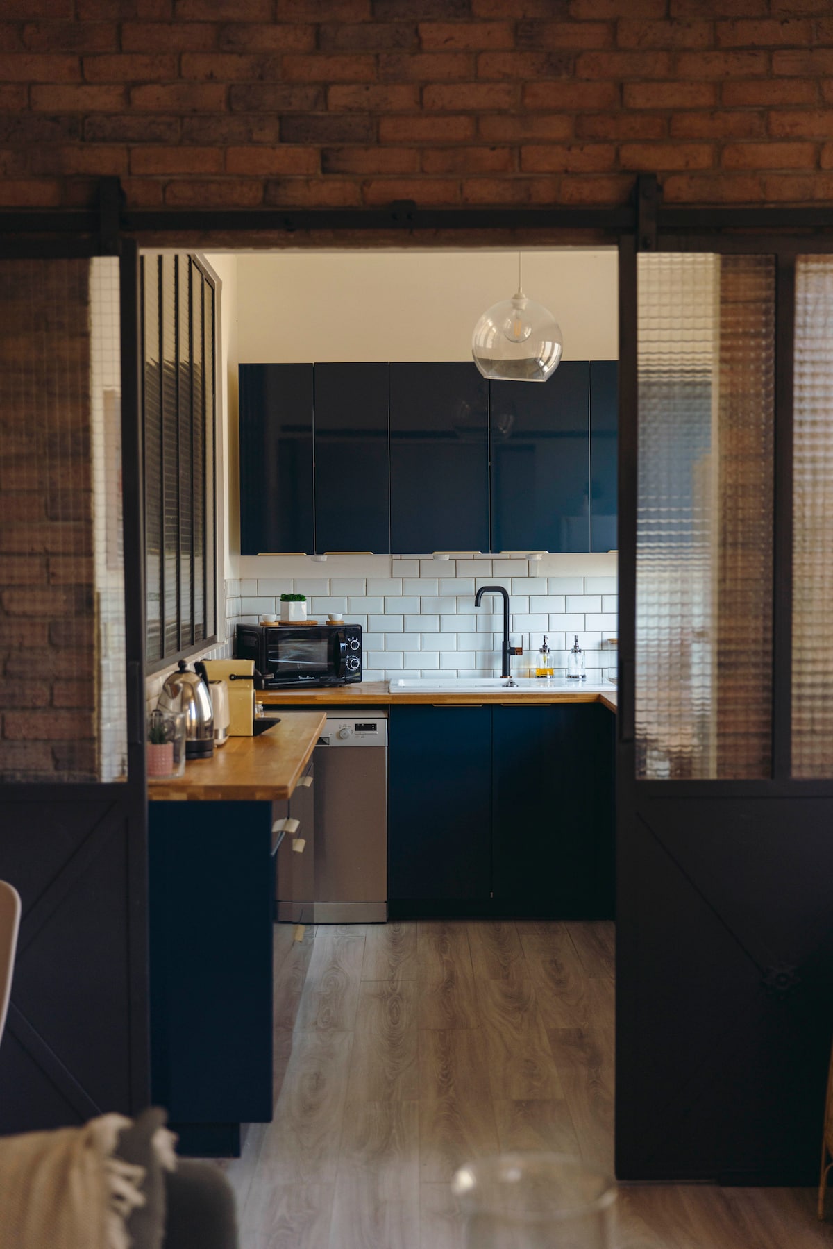 A fully equipped kitchen is situated behind a glass door, featuring dark cabinetry and a wooden countertop. The backsplash is lined with white tiles, complemented by modern appliances, including a microwave and a dishwasher. Light is provided by a hanging pendant lamp.