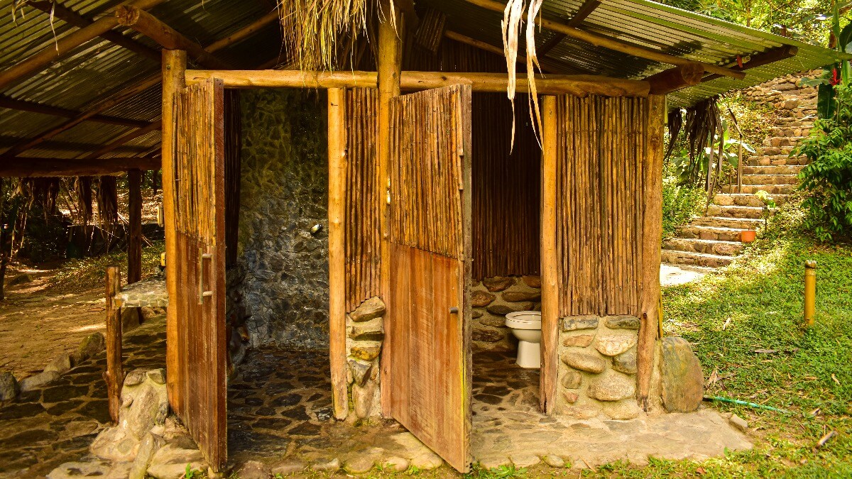 A rustic outdoor bathroom is framed by natural wooden beams and bamboo, with stone walls and a stone floor. Open wooden doors reveal a toilet and space for personal care. Lush greenery and a pathway lead to a staircase in the background.