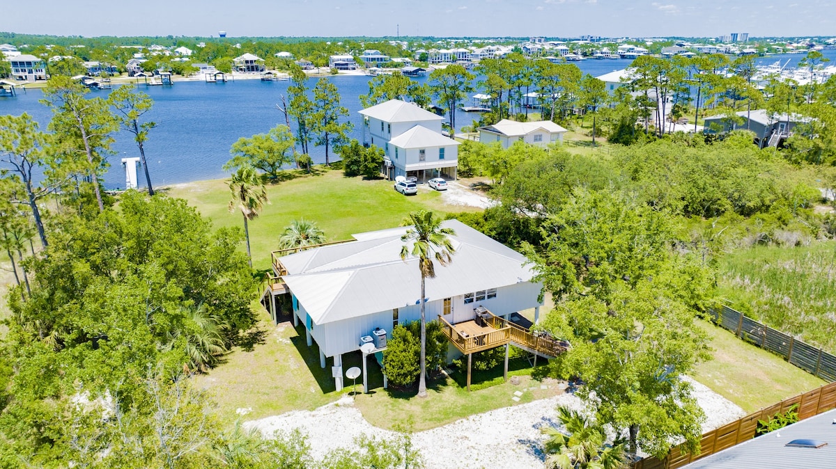 An aerial view showcases a charming single-story home surrounded by greenery and water. The property features a large deck with seating, bordered by lush trees and shrubs, and offers glimpses of nearby houses and boats in the water.