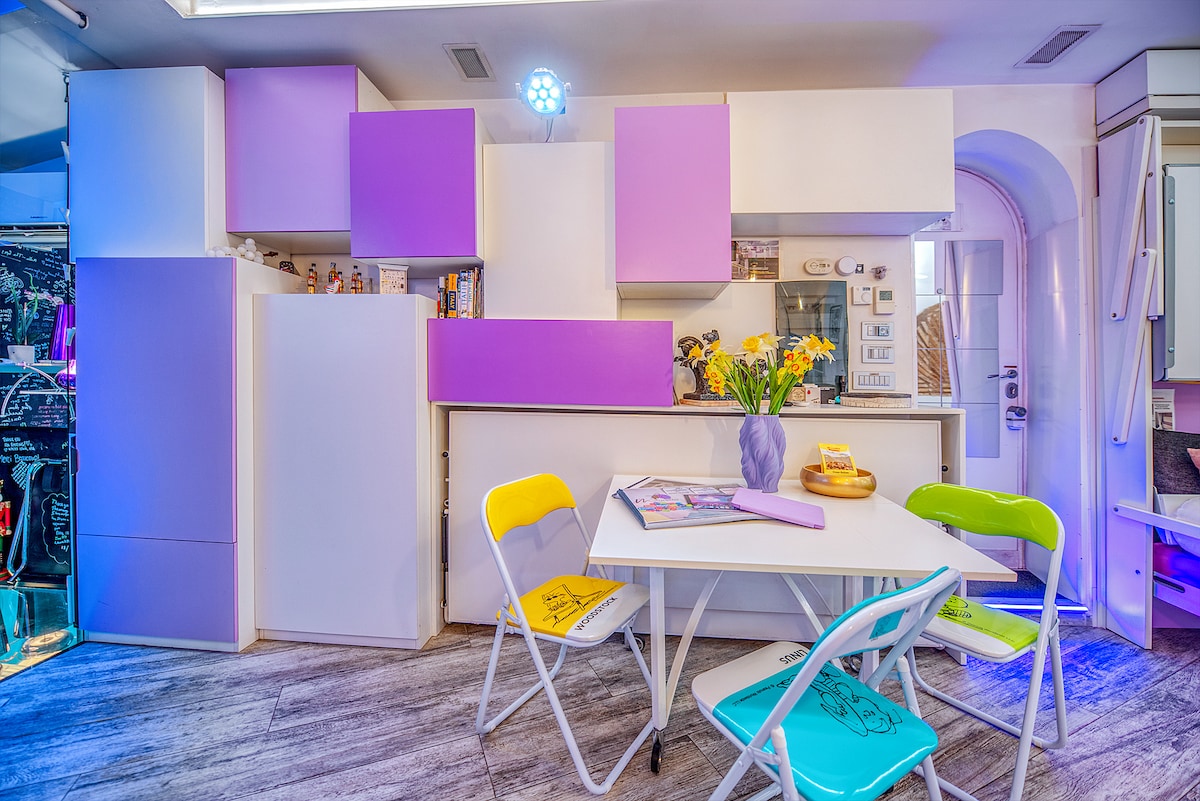 A modern kitchen area features a mix of white cabinetry with purple accents, creating a vibrant atmosphere. A small white table, surrounded by colorful chairs in yellow, green, and blue, is inviting for meals or activities. Natural light fills the space, enhancing its cheerful design.
