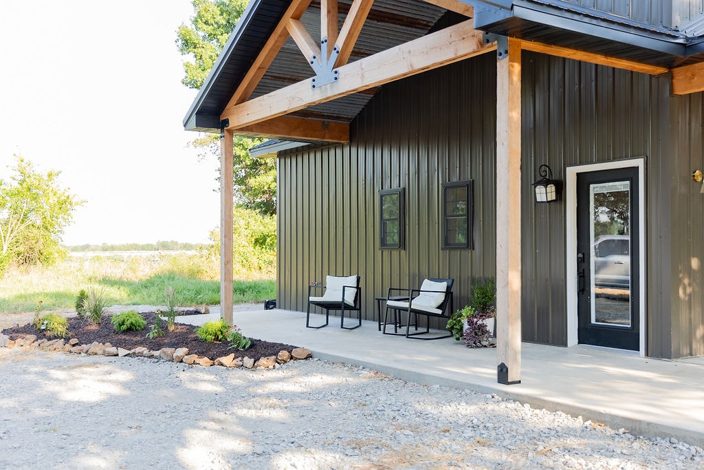The exterior space features a covered porch with two chairs positioned on a smooth concrete surface. Green landscaping, including small plants and rocks, extends along the edge, with open fields visible in the background. The entrance showcases a black door and large windows.