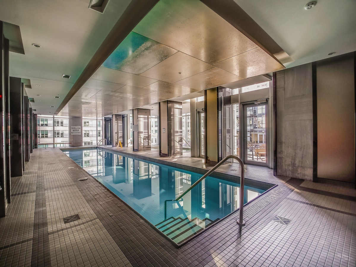 The indoor pool area features a sleek design with large windows allowing natural light. Tile flooring surrounds the swimming area, which is complemented by modern seating options. The space maintains a clean and inviting atmosphere, perfect for relaxation.