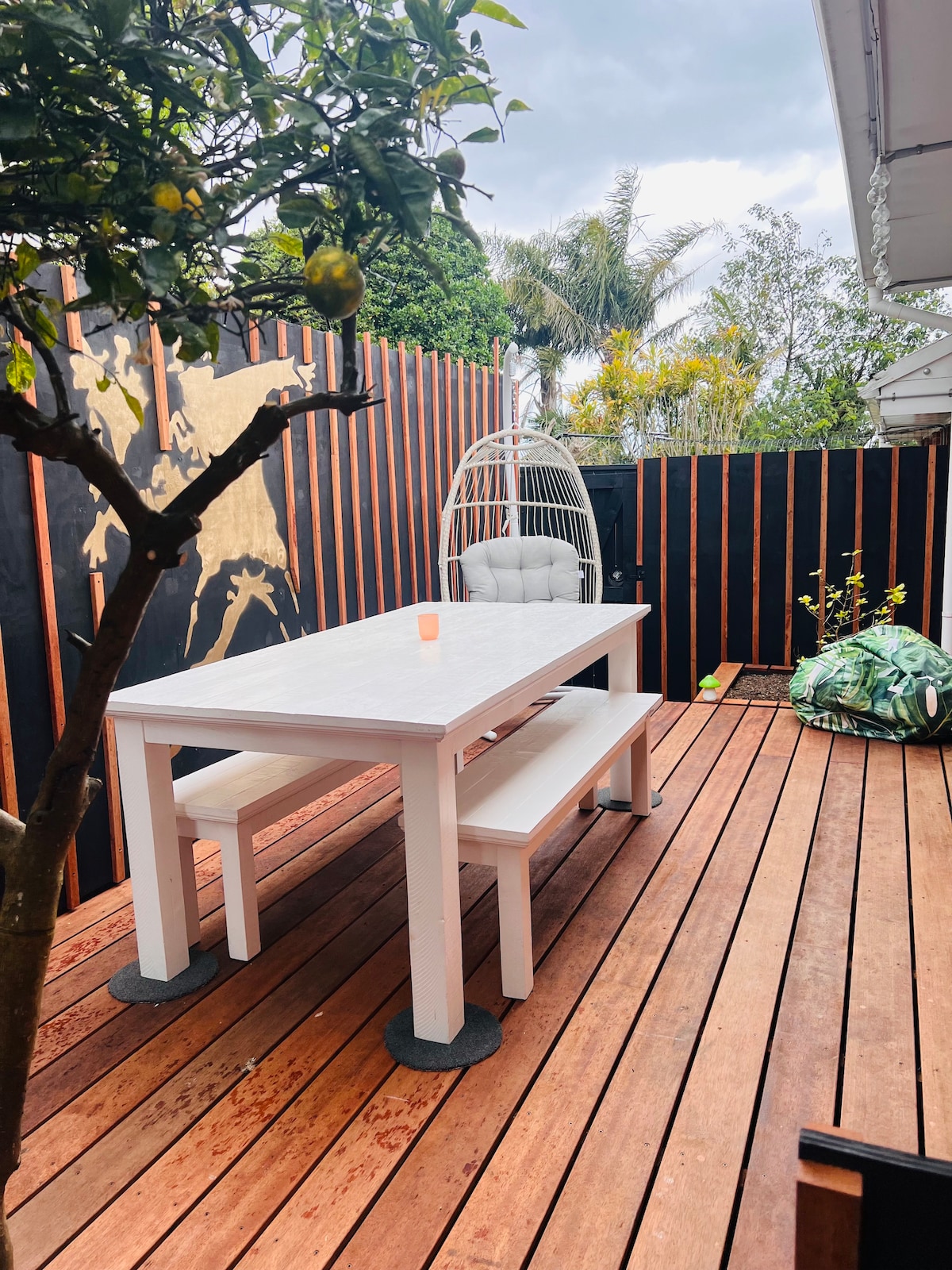An outdoor patio area features a wooden deck with a white dining table and two benches. A cozy hanging chair is positioned nearby, surrounded by greenery. Decorative elements include a lemon tree and a colorful beanbag, creating a relaxing space for outdoor enjoyment.