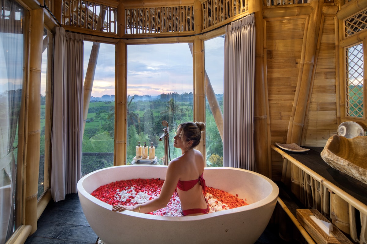 A large circular bathtub is filled with floating petals, surrounded by a wooden structure featuring large windows. Soft natural light filters through sheer curtains, illuminating the tranquil scene. Candles on a nearby table add a serene touch to the relaxation area with scenic views outside.