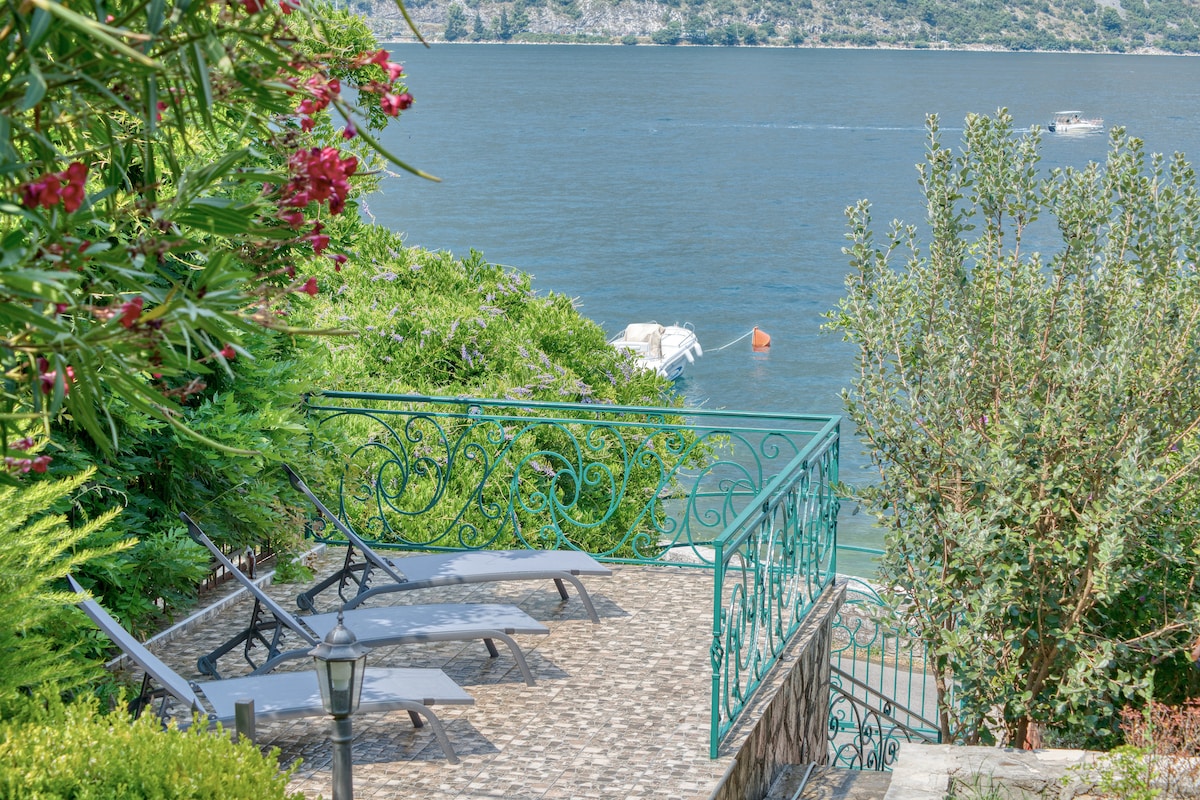 A stone terrace opens to views of Kotor Bay, featuring a decorative wrought-iron railing. Several lounge chairs are arranged for relaxation, surrounded by lush greenery and vibrant flowers. The calm water is visible in the background, enhancing the serene setting.