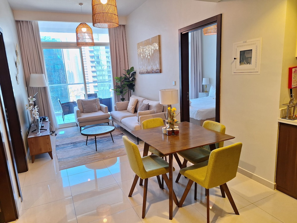An open-plan living area features a comfortable sofa and a round coffee table, positioned on a stylish area rug. A dining table with yellow chairs is visible, alongside large windows that offer natural light and views of the balcony. The space is bright and inviting.