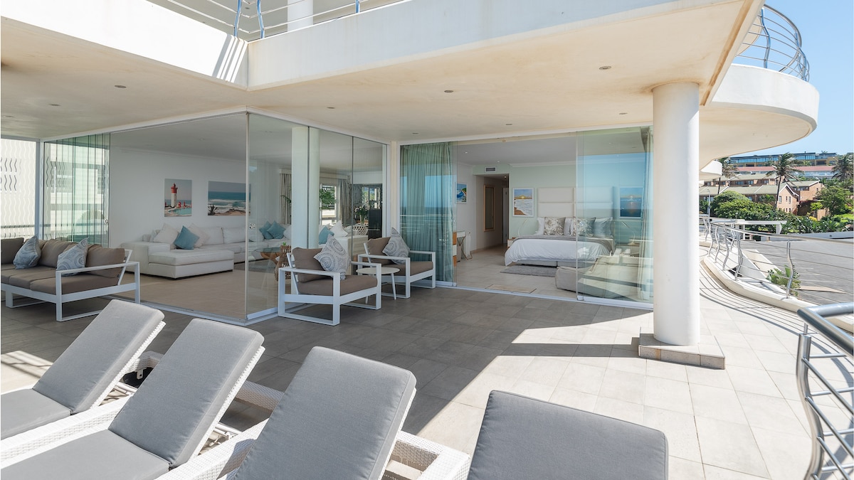 An expansive terrace features comfortable lounge chairs, providing ample space for relaxation. Large glass doors offer a view into the bright interior space, furnished with light-colored decor, creating a seamless connection between outdoor and indoor areas. A serene coastal ambiance is evident.