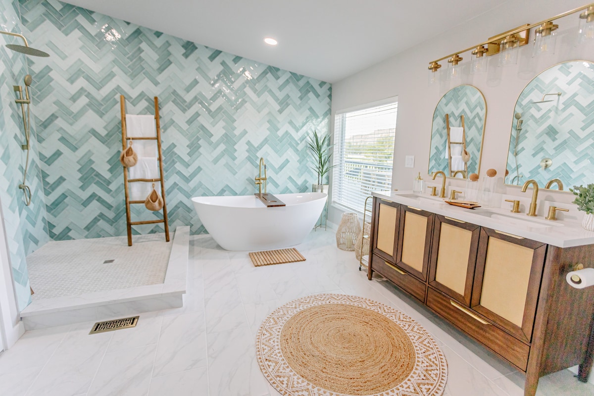A modern bathroom features a soaking tub complemented by a stylish faucet. A spacious shower with a glass door is visible, alongside a double vanity with gold fixtures. Decorative tiles in a herringbone pattern adorn the walls, while natural light filters through a window.