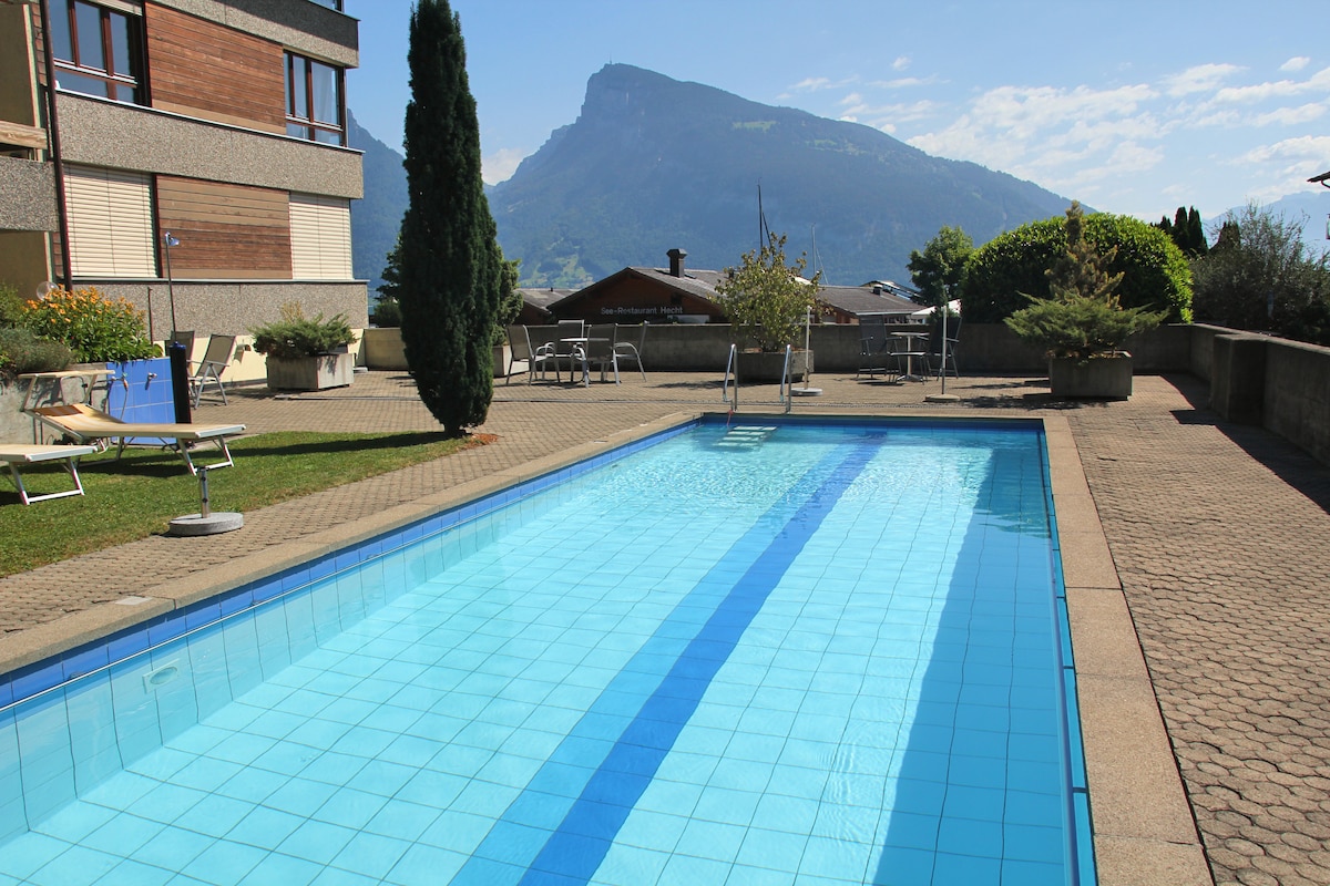 A clean outdoor pool is surrounded by a paved area, with lounge chairs positioned nearby. Lush greenery and planters add natural elements to the setting. The backdrop features a mountain range under a clear blue sky, enhancing the serene atmosphere of the space.