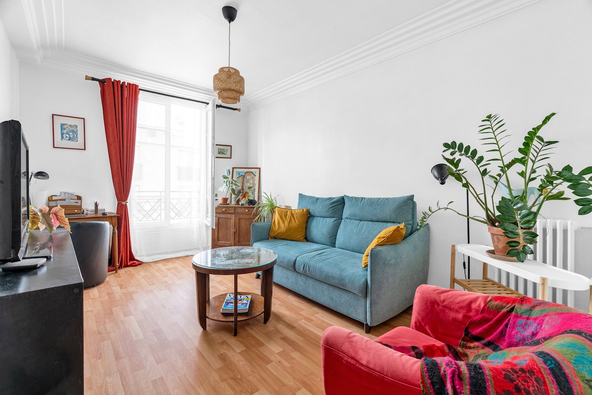 A bright living area is presented, featuring a comfortable blue sofa alongside a vibrant armchair. A glass coffee table occupies the center, with decorative plants adding a touch of green. Natural light floods in through the window, framed by red curtains that enhance the space.