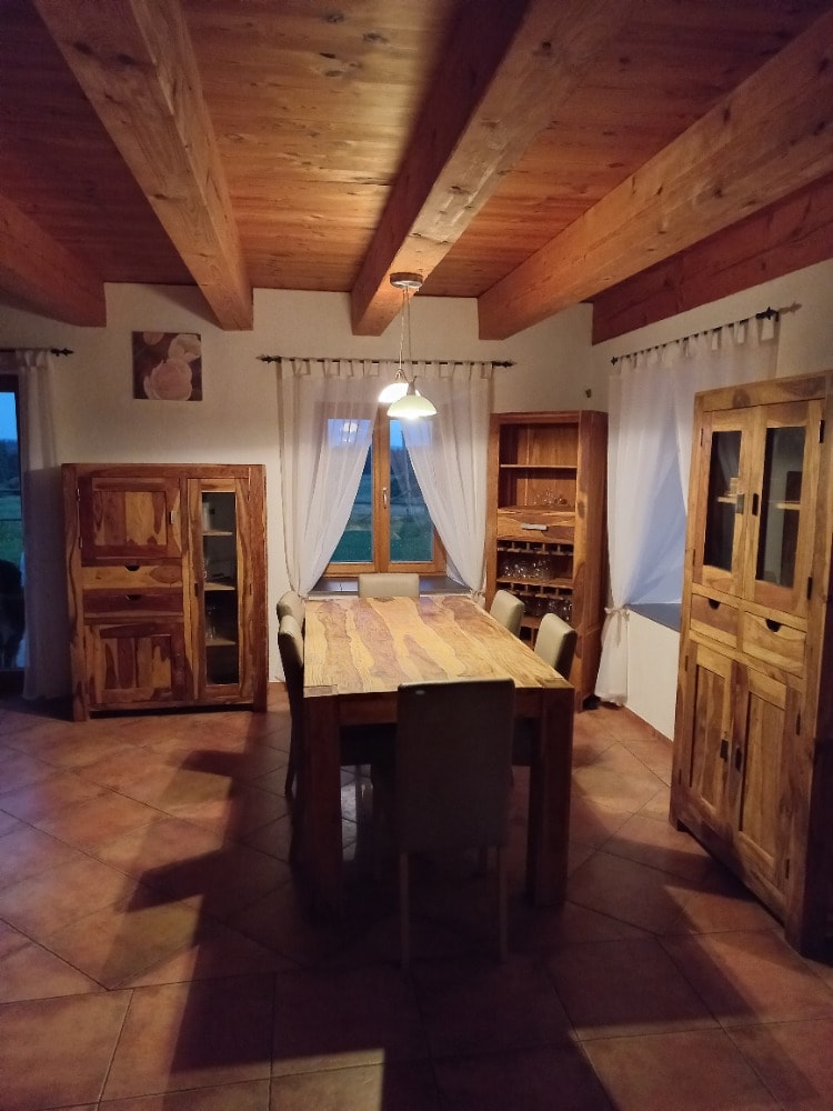 The dining area features a large wooden table surrounded by chairs, illuminated by a pendant light. Natural light enters through a window, and wooden beams enhance the room's character. A storage cabinet and another cupboard are positioned against the walls.