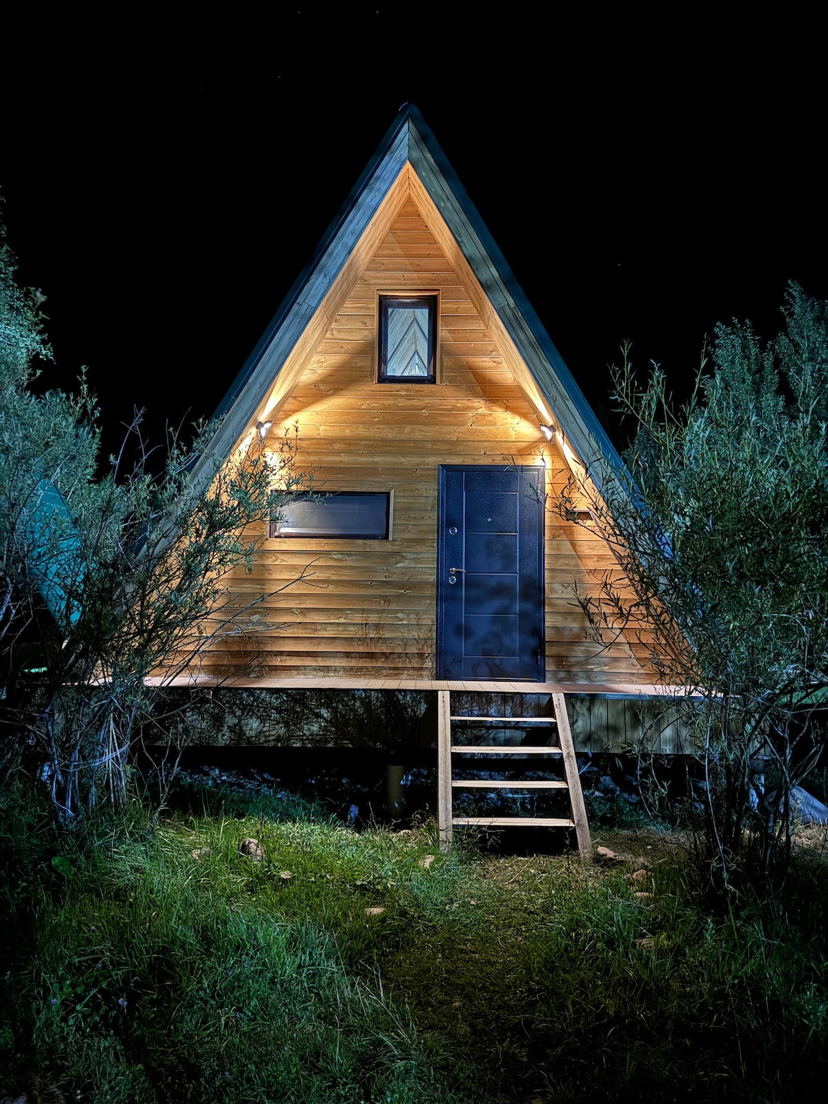 A triangular A-frame cabin is illuminated at night, showcasing its wooden exterior and distinct features. A set of steps leads to the door, while shrubs frame the structure, emphasizing the cabin's connection with the surrounding nature.