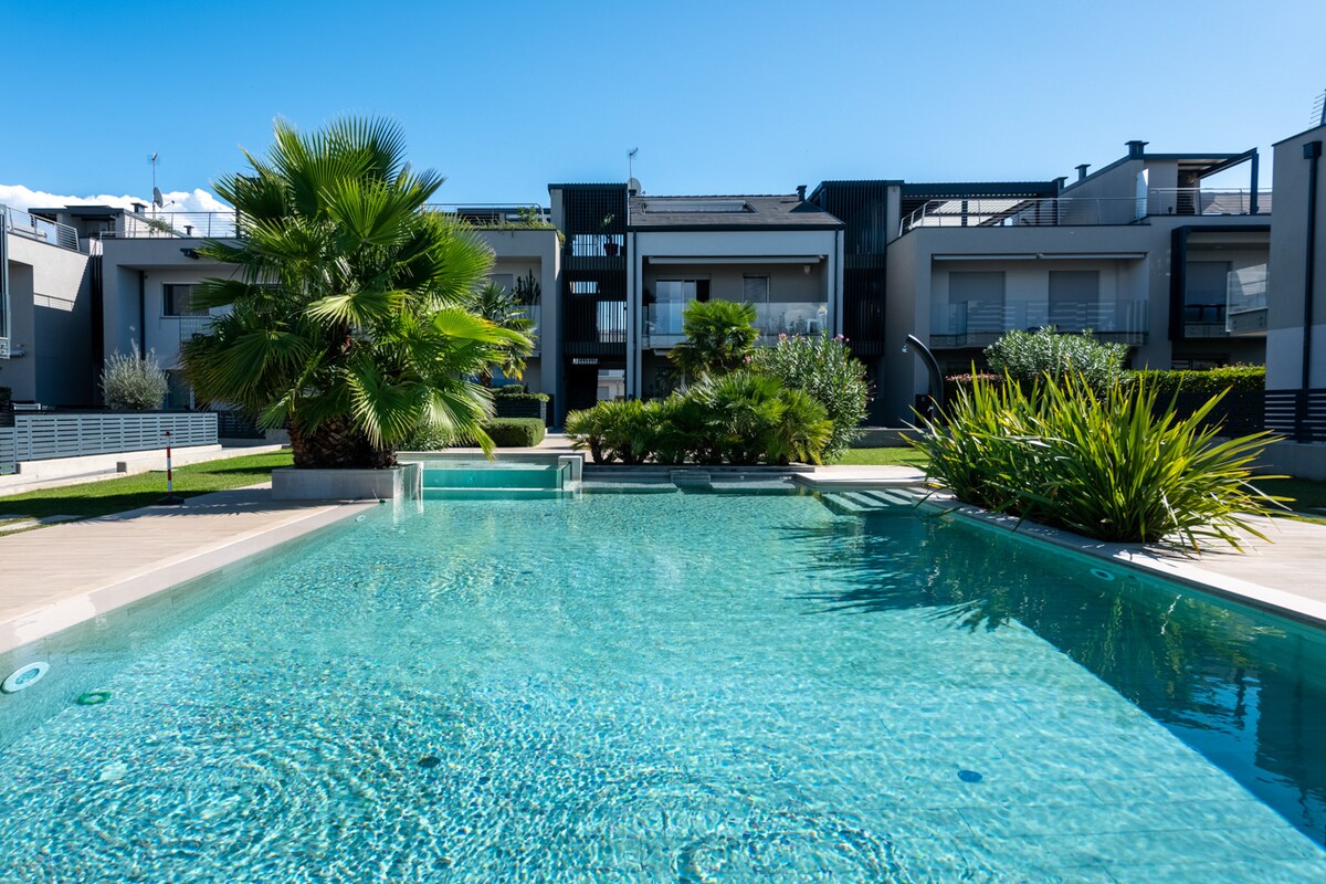 A clear swimming pool surrounded by well-maintained greenery and palm trees is visible. The tranquil water reflects the blue sky, while the modern residence is seen in the background, enhancing the outdoor space's relaxing atmosphere.