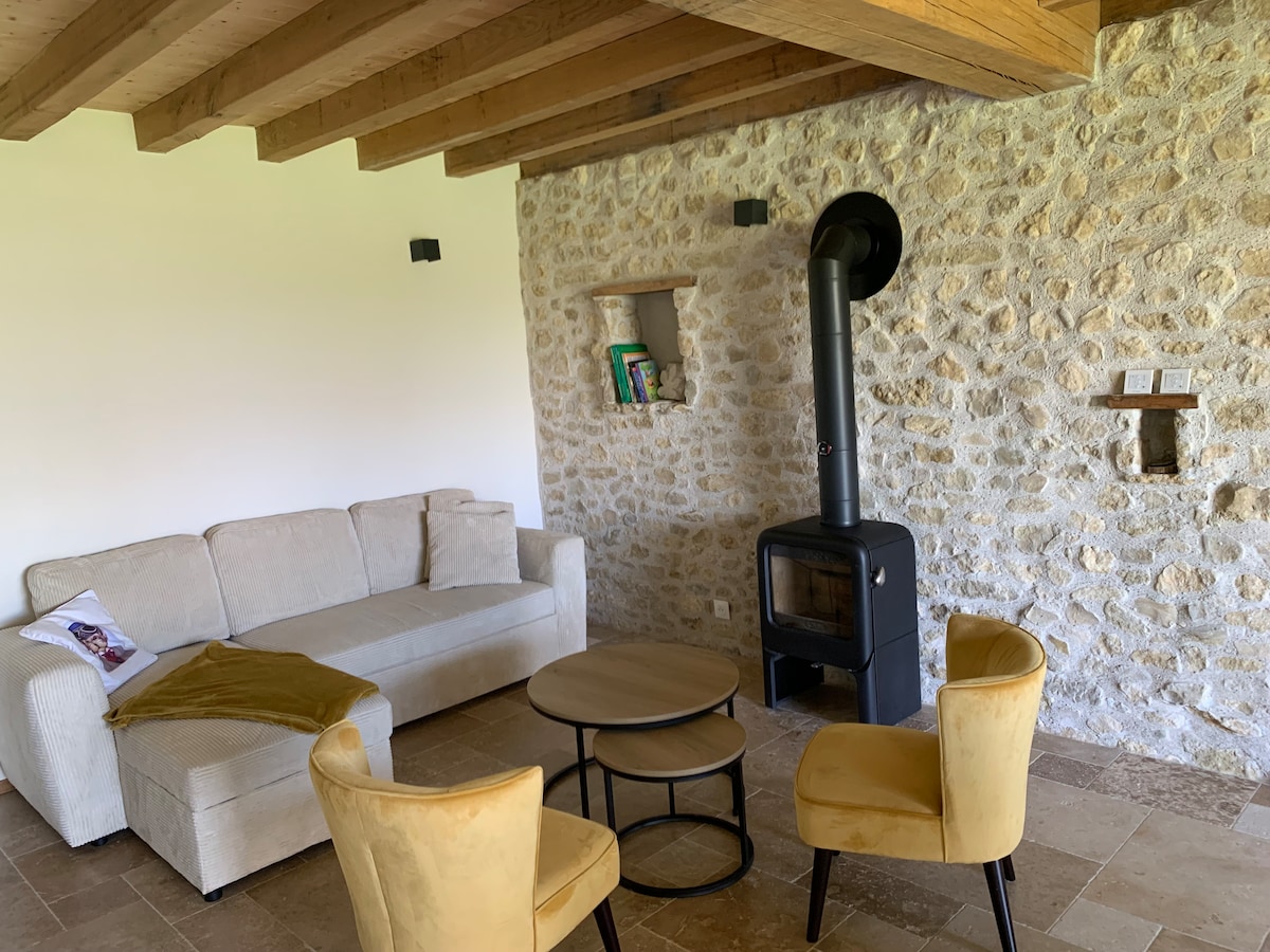 A welcoming living area is presented with a neutral-colored sofa and two yellow chairs positioned around a circular table. A wood-burning stove is integrated into a stone wall, while shelves display books. Exposed wooden beams stretch across the ceiling, contributing to the room's character.