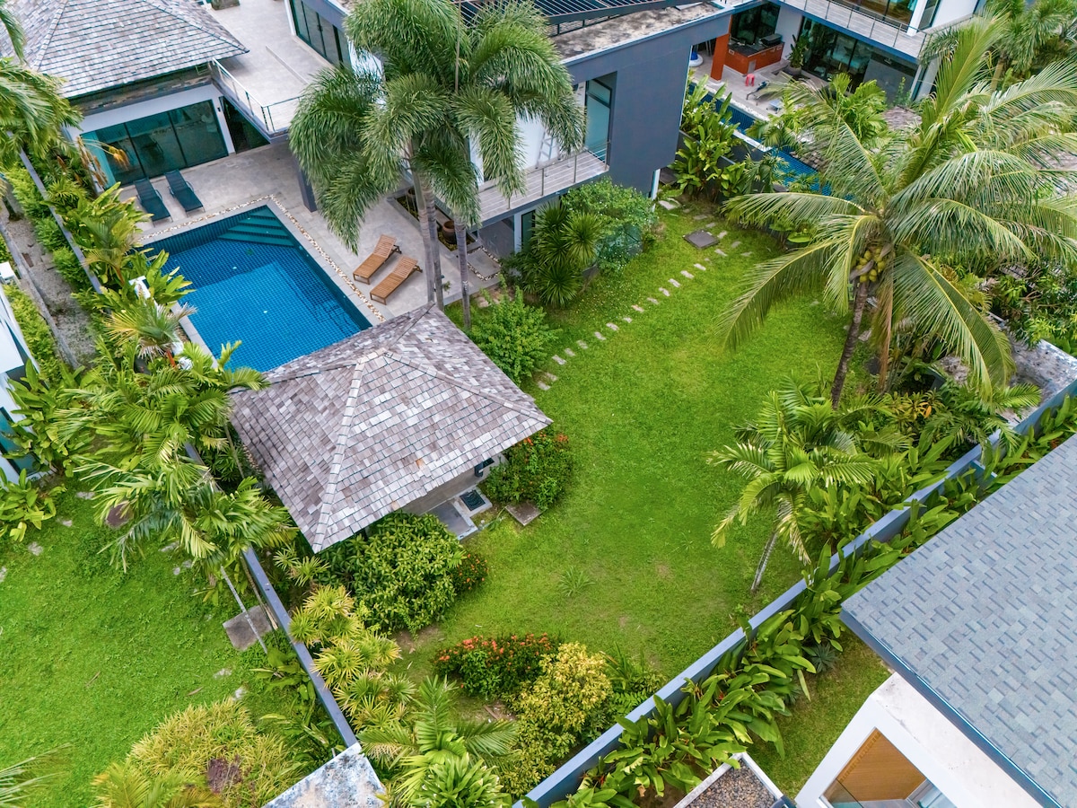An aerial view captures the lush tropical garden surrounding the villa, featuring manicured green lawns and various palm trees. A thatched gazebo adds character, while a spacious swimming pool glimmers nearby, complemented by lounge chairs under the shade of trees.