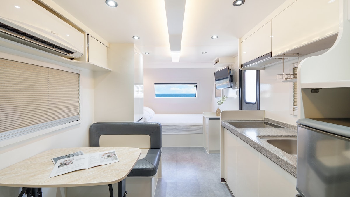 An interior view of a modern caravan displays a compact layout with a kitchenette, dining area, and sleeping space visible in the background. Natural light enters through a large window, highlighting the sleek cabinetry and neutral color palette throughout the space.