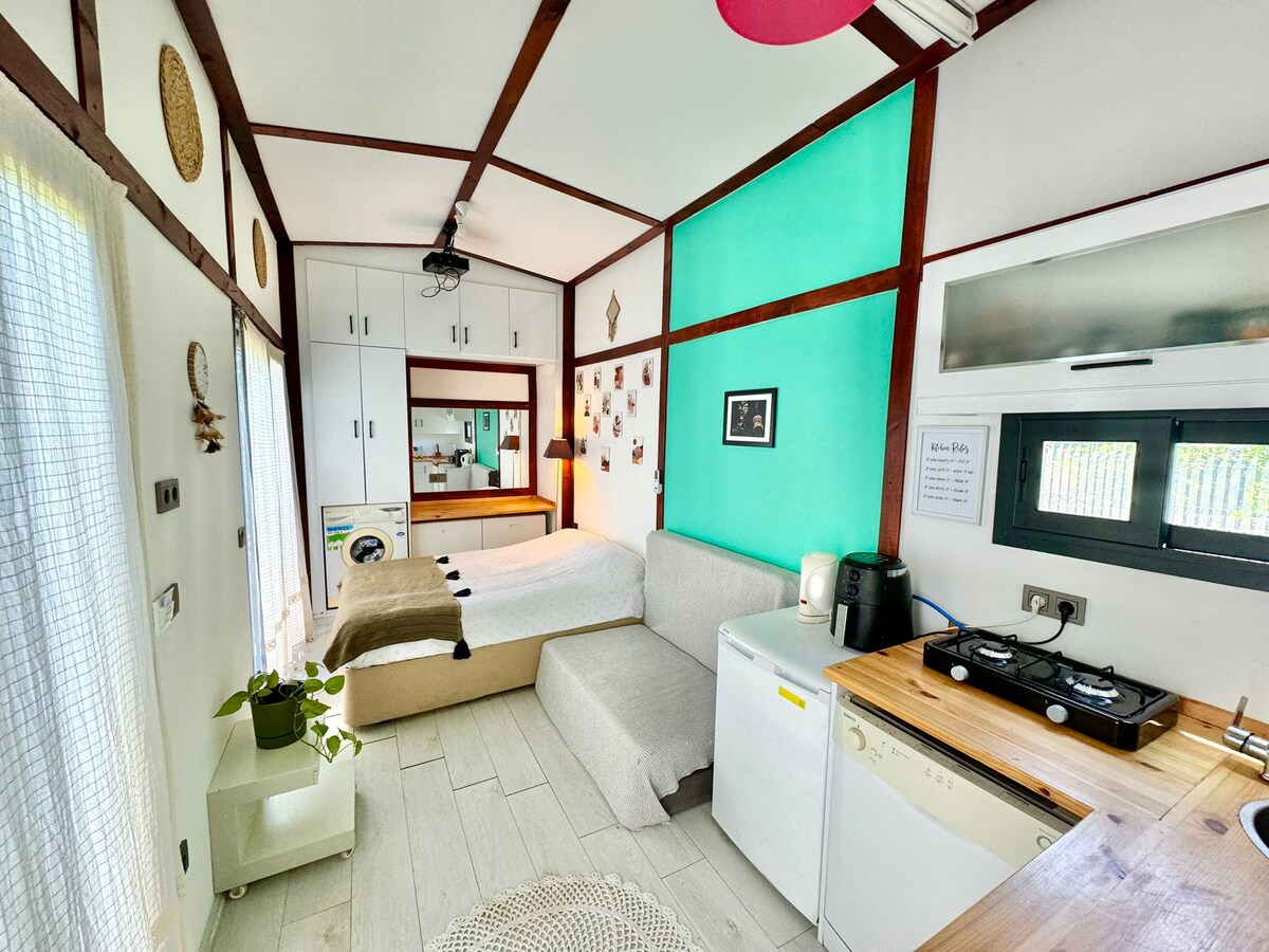 A well-organized interior space features a spacious bed accompanied by a versatile sofa that can accommodate additional guests. A kitchenette with essential appliances, including a compact refrigerator and a stove, offers convenience. Natural light enters through a large window, enhancing the bright, airy atmosphere.