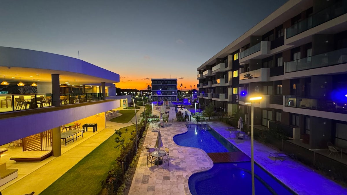 The image depicts a modern condominium complex at dusk, featuring silhouette outlines of buildings against a colorful sunset sky. A pool area with smooth stone tiles is visible, alongside greenery and lounge seating, creating an inviting atmosphere for relaxation.