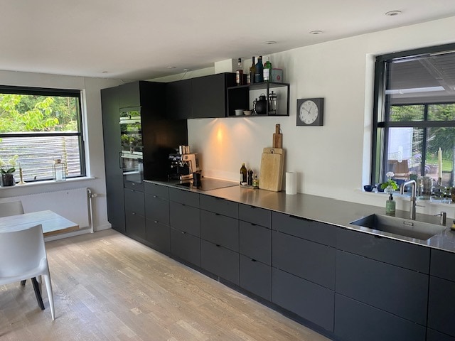 A modern kitchen is presented with sleek black cabinets and a minimalist design. A neutral countertop accompanies a built-in espresso machine and various kitchen appliances. Natural light filters through large windows, illuminating the space and providing a view of the surrounding greenery.