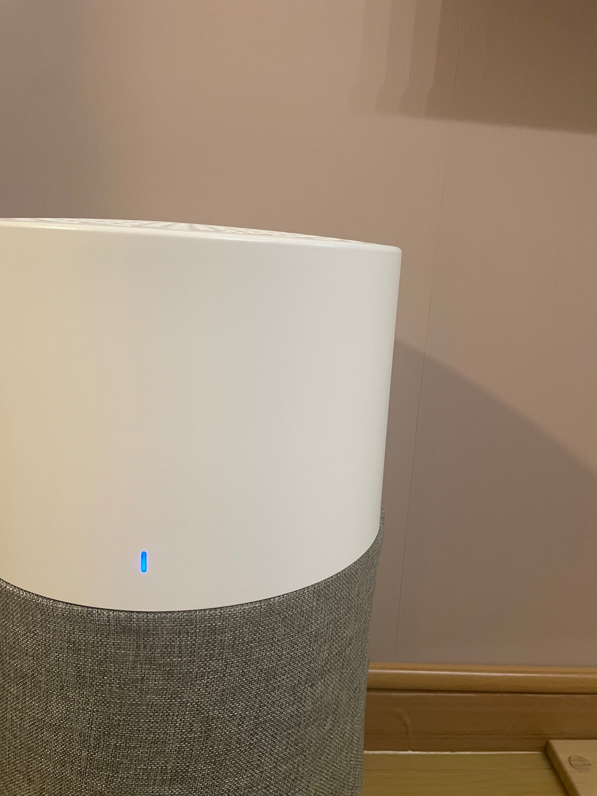 A modern air purifier is positioned in the corner of a room, featuring a sleek white top section and a textured gray fabric base. A soft blue light is visible on the front, indicating the device is powered on.