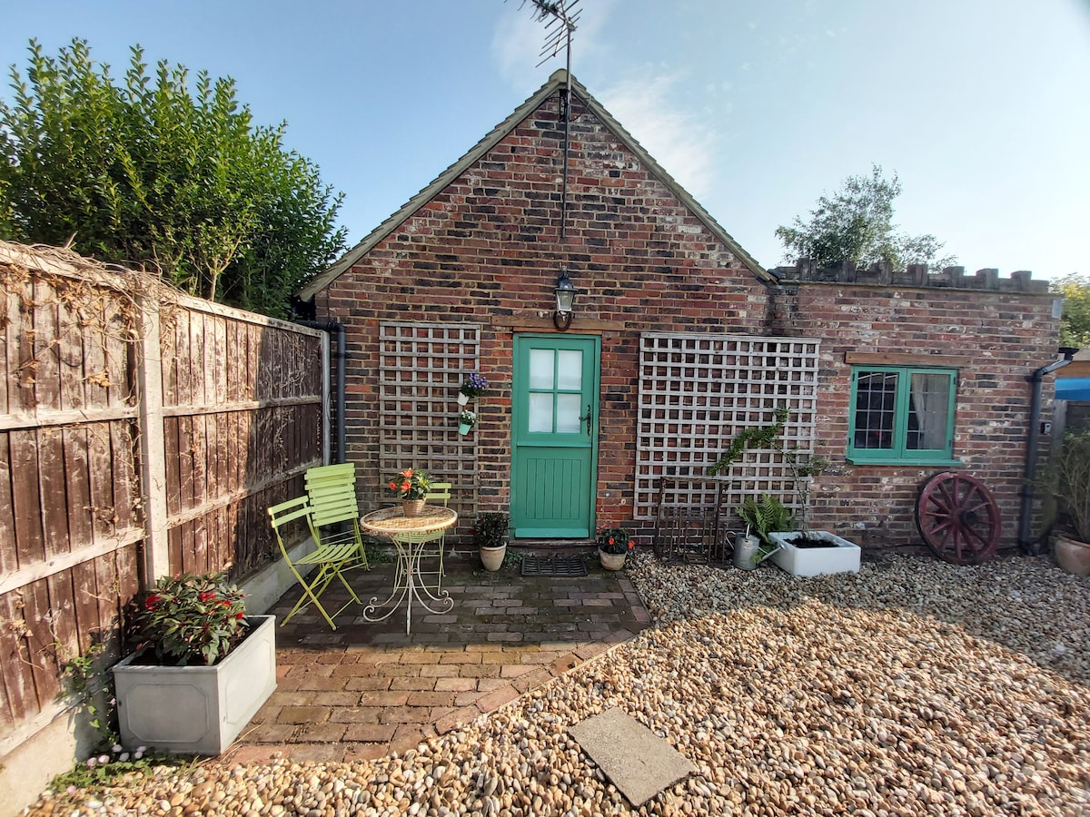 The exterior of The Old Coach House features a charming brick facade with a green door, surrounded by a gravel area. Two green chairs and a small table are positioned outdoors, with potted plants nearby, creating a pleasant, inviting entrance.