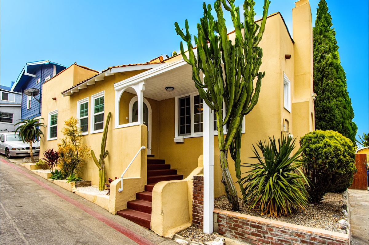 A charming beach bungalow exterior is depicted, featuring stucco walls painted in warm tones. Lush desert plants and cacti accentuate the landscape. The home's inviting entrance is highlighted by a white railing and steps leading to the front door, framed by large windows that allow natural light.