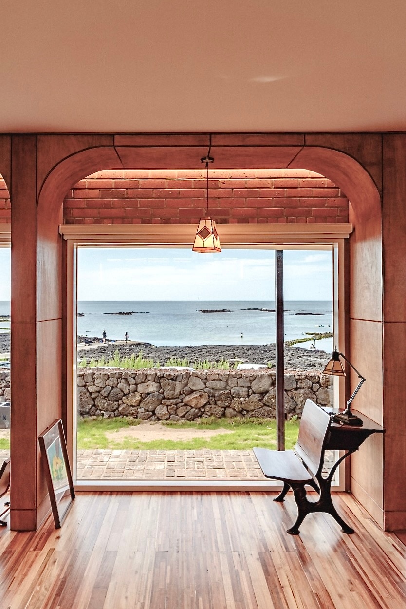 A view through large glass doors reveals a tranquil ocean scene, framed by inviting wooden finishes. A small wooden desk with a lamp sits in the foreground, enhancing the cozy aesthetic. Natural light illuminates the warm wooden flooring, accentuating the connection between indoor and outdoor spaces.