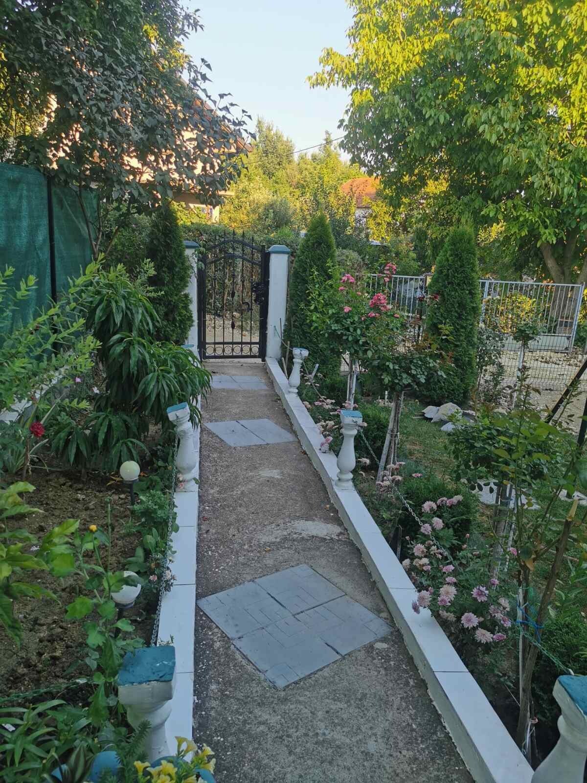 A well-maintained garden path is framed by lush greenery and colorful flowerbeds. Decorative stone borders line the walkway, leading towards a wrought-iron gate that opens to the exterior. Tall shrubs and various ornamental plants enhance the inviting atmosphere.