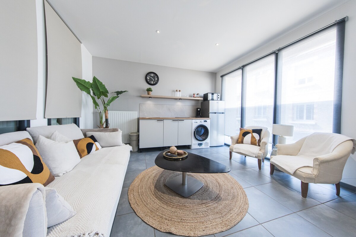 A bright living area features a comfortable sofa adorned with patterned cushions and an elegant coffee table at its center. Large windows invite natural light, and a small indoor plant adds a touch of greenery. A washing machine is discreetly positioned alongside the modern kitchen setup.