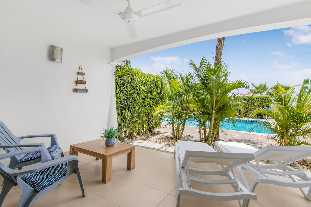 A private terrace features a wooden table and seating area with lounge chairs, surrounded by tropical plants. The terrace overlooks a swimming pool, providing a bright and airy atmosphere with clear skies in the background.