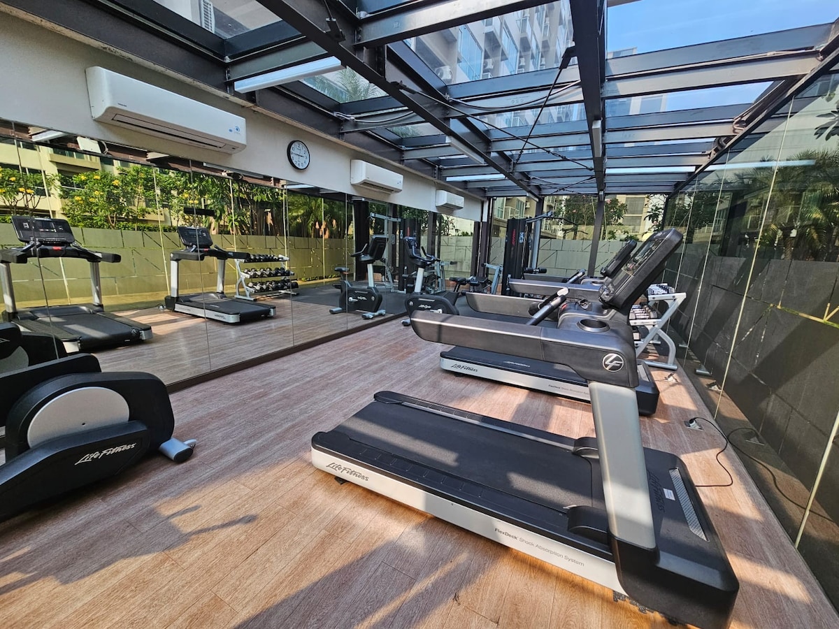 A fitness area is equipped with modern exercise machines, including treadmills and stationary bikes. Floor-to-ceiling windows provide natural light while showcasing greenery outside. The space is complemented by wooden flooring and mirrors that enhance the area's openness.