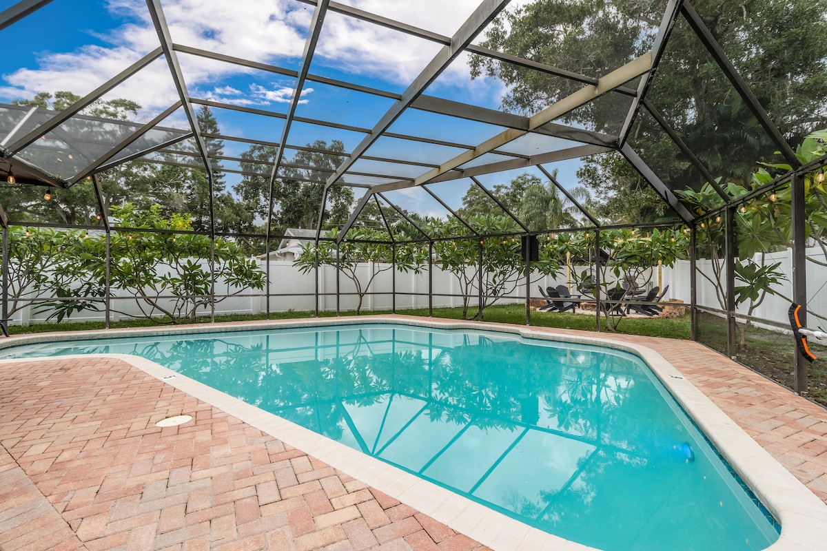 A spacious screened-in enclosure surrounds a refreshing pool with clear water, softly reflecting the sky above. Lush greenery and landscaped shrubs provide privacy, while a patio area with seating is visible in the background.