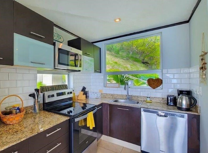 A modern kitchen is equipped with stainless steel appliances, including a stove, refrigerator, and dishwasher. A deep sink is positioned under a large window that offers views of the surrounding greenery. Granite countertops provide ample workspace, complemented by dark cabinetry and light-colored backsplash tiles.