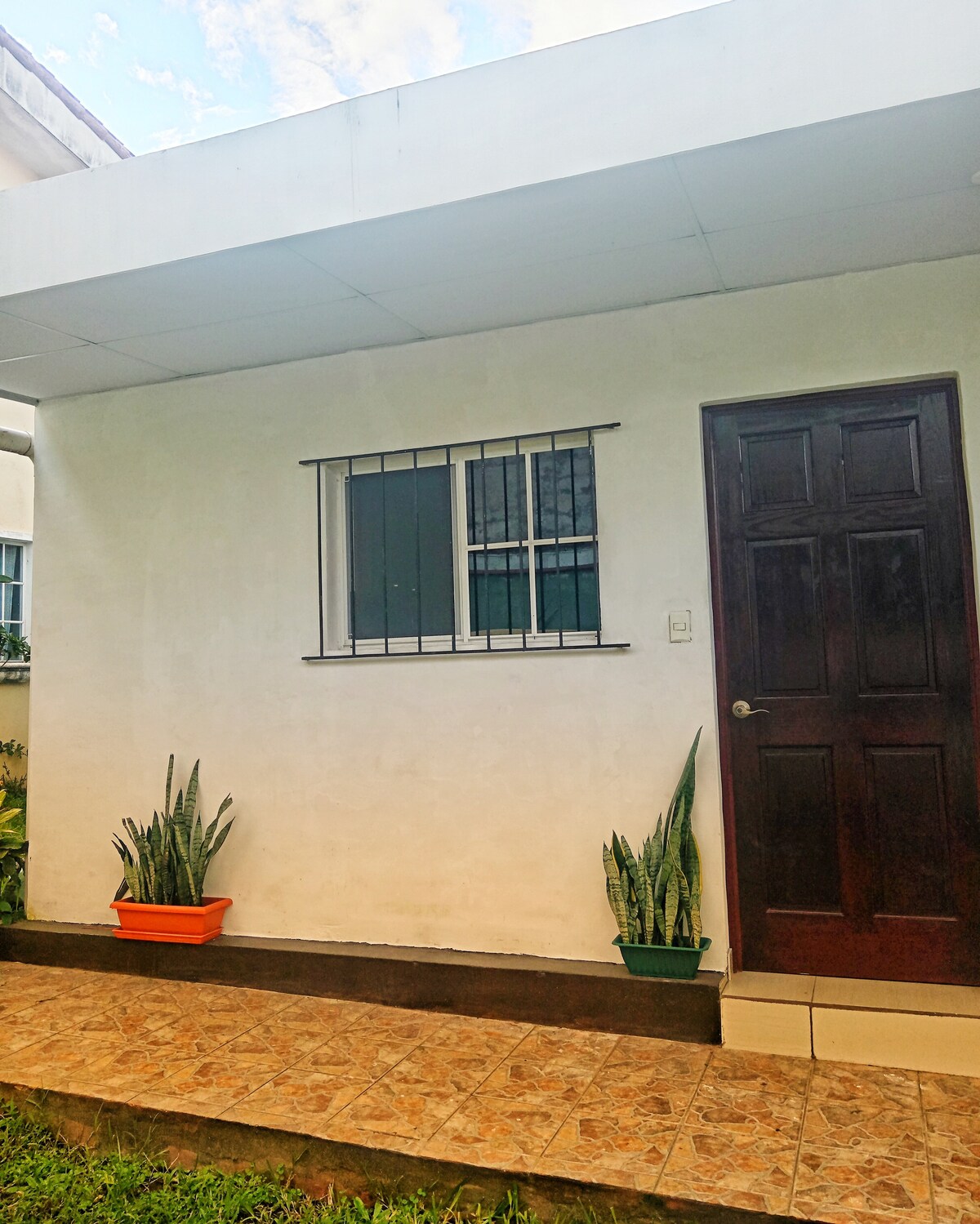 An exterior view of a small, independent entrance is visible, featuring a dark wooden door and a window with metal grilles. Two potted plants are positioned on either side of the entrance, enhancing the welcoming appearance. The walkway is paved and bordered by green grass.