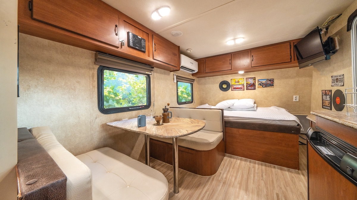 A cozy interior of a caravan features wooden cabinetry and a beige color scheme. A dining area with a light-colored table and comfortable seating is positioned across from a spacious sleeping area, which includes a queen bed. Natural light filters through the windows, revealing a green outdoor view.