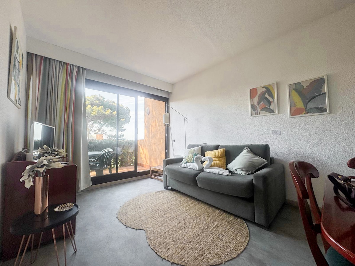 A cozy living area is shown, featuring a comfortable grey sofa with decorative cushions. A small round rug is placed on the floor. Large sliding glass doors lead to a private terrace, allowing natural light to fill the space, while framed artwork decorates the walls.